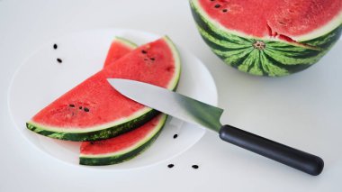 Close-up slices of fresh ripe red watermelon on a plate with a knife on the table on a white background, half of a watermelon. Ripe juicy summer berries, melon season. Macro photo.