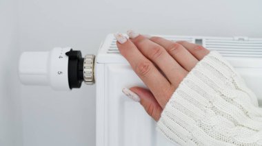 Close-up of a woman in white sweater warms her hands over the battery on background of white wall.Macro photo of woman's hands,copy space.Concept of saving gas in winter.Gas crisis in Europe