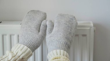Close-up of woman in white sweater and gray mittens warms her hands over a radiator against a white wall. Macro photo of female hands, copy space. Gas saving concept in winter. Gas crisis in Europe