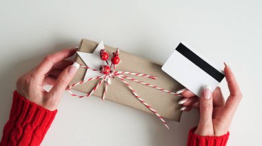 Kırmızı kazaklı kadının elleri elinde kredi kartı ve hediye kutusuyla beyaz arka plan, üst görüş, daire, fotokopi odası. Noel ve yeni yıl alışverişi, kredi kartı ödemesi. İnternetten hediyeler alıyor.