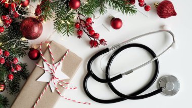 Noel medikal afişi. Hediye kutusunu, köknar dallarını, konileri, köknar ağaçlarını, kırmızı topları ve steteskopu beyaz arka planda, üst tarafta, düz yerde, kopyalama alanında kapatın. Yeni yıl tıbbı, doktor için tebrikler.
