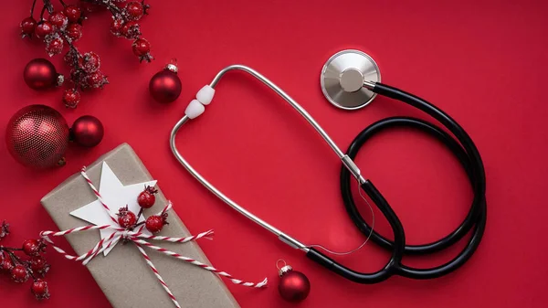Christmas medical banner. Close up gift box, berries, red balloons and stethoscope on red background, top view, flat lay, copy space. New Year\'s medicine, congratulations for the doctor