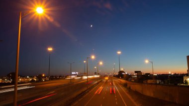Long exposure shot of busy traffic on a highway under the moon and blue and orange sunset sky in Toronto, Ontario, Canada. 