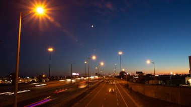 Long exposure shot of busy traffic on a highway under the moon and blue and orange sunset sky in Toronto, Ontario, Canada. 