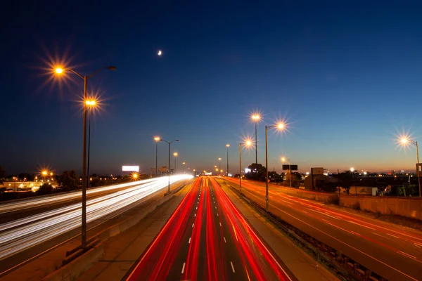 Long exposure shot of busy traffic on a highway under the moon and blue and orange sunset sky in Toronto, Ontario, Canada.