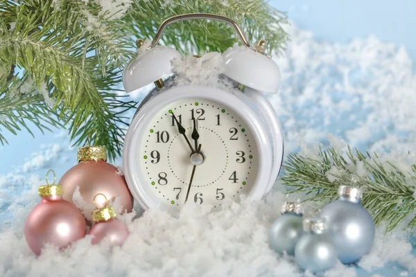 The white clock shows the time 23-55. The last minutes of the outgoing year. The alarm clock stands in the snow on a blue background with New Years decorations, fir branches and Christmas balls. — Stock Photo, Image