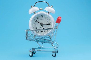 Mavi arka planda süpermarket tramvayındaki beyaz saat. Konsept - bir çevrimiçi mağazadan mal alarak satın almaktan tasarruf etmek, coronavirüs nedeniyle uzaktan satın almalar.