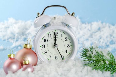 Beyaz saat 23-55 'i gösteriyor. Giden yılın son dakikaları. Çalar saat, kar üzerinde pembe Noel toplarıyla mavi bir zemin üzerinde duruyor. Yeni yıl kartı, yatay fotoğraf