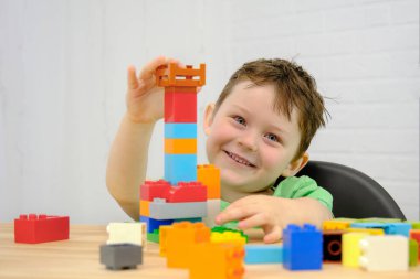 Mutlu çocuk bir inşaatçıyla oynuyor. Çocuk 5 yaşında, beyaz tenli. Masada oturuyor ve kameraya bakıyor, gülümsüyor. Masanın üzerinde renkli küpler, oyuncaklar var. Yatay fotoğraf.