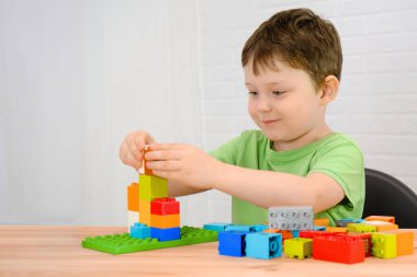 Yeşil tişörtlü bir çocuk odadaki bir masaya oturur ve renkli küplerden bir müteahhit alır. Çocuk gülümsüyor, kameraya bakmıyor. Yatay fotoğraf. Oyuna tutkulu.