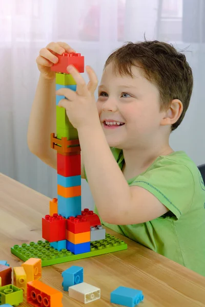 Ein glückliches Kind sammelt einen Turm aus farbigen Würfeln. Die Vorschulkinder setzen sich an den Tisch und bauen das Bauset zusammen. vertikales Foto. Der Junge lächelt, zufrieden mit dem Ergebnis, — Stockfoto