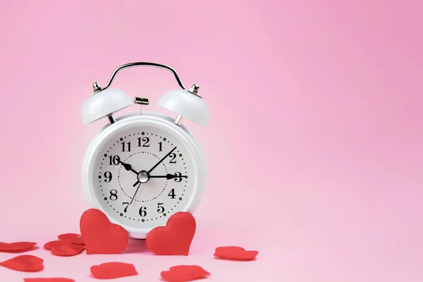 White alarm clock stands on a pink background among red hearts. The idea is that its time to shop for Valentines Day. Horizontal photo. Imagen de archivo