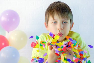 Çocuk ellerinde çok renkli bir konfeti patlatıyor. Beyaz bir duvar ve balonlara karşı sarı tişörtlü bir çocuk. Kafkasyalı bir çocuğun portresi, 7-8 yaşlarında. Konsept - Mutlu yıllar, parti, tatil
