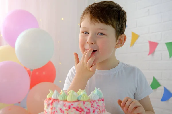 お祝いの席の少年は指でケーキを試してみる。指を口の中に。子供は、笑顔、満足し、幸せそうに見えます。風船や旗を背景に4 〜 5歳の少年。. — ストック写真