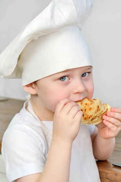 Een jongen met blauwe ogen van 4-5 jaar oud eet een pannenkoek. hij is in de vorm van een kok, in een witte dop en schort. Verticale foto. Het kind kijkt in de camera. Proef een zelfbereide schotel — Stockfoto