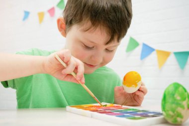 4-5 yaşlarında bir çocuğun portresi. Yeşil tişörtlü bir çocuk sarı boyalı ahşap bir Paskalya yumurtası boyuyor. Elinde bir yumurta ve fırça tutuyor. Yaratıcı uğraş, Paskalya hazırlığı.