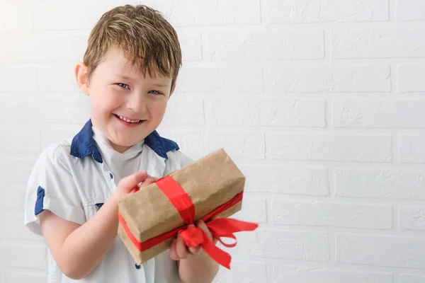 Porträt eines glücklichen Jungen in weißem Hemd mit einem Geschenk in der Hand. Das Kind lächelt, hält eine Schachtel mit einer roten Schleife in den Händen. — Stockfoto