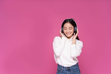 Pembe arka planda müzik dinlerken ve dans ederken kulaklık takan güzel, gülümseyen Asyalı bir kadın.