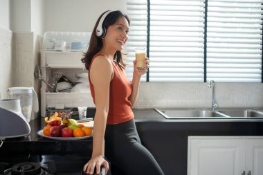 Güzel genç bir kadın, kulaklıkla müzik dinlerken evdeki meyvelerden meyve suyu yapıyor ve içiyor. Yaşam tarzı kavramları.