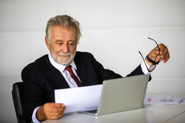 An elderly businessman sitting at his desk checking documents with a notebook computer on his desk.