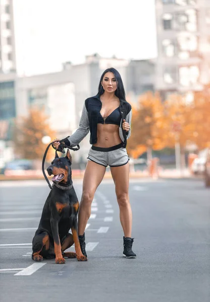 Beautiful fitness brunette girl with perfect shapes walking on the city square in sexy sports costume with huge Doberman dog in the rays of setting sun.