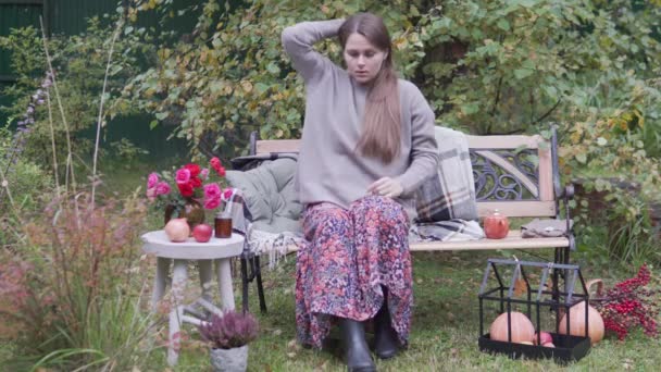 Piękna młoda kobieta w długiej sukience siedzi na ławce w jesiennym ogrodzie przy stole z wazonem kwiatów — Wideo stockowe