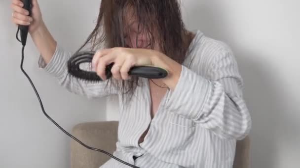 Eine Frau im Nachthemd trocknet dunkle Haare mit einem Haartrockner, nachdem sie zu Hause auf einem Stuhl geduscht hat — Stockvideo