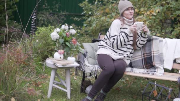 Egy gyönyörű fiatal nő egy csésze forró itallal ül az őszi parkban egy padon az asztalnál virágokkal. — Stock videók