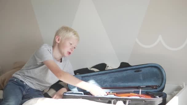 The child does not want to do violin at home, the boy sits on the bed by the open cover with the violin — Stock Video