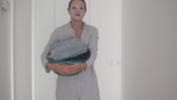 Een grappige vrouw met een waskom van gewassen kleren in haar handen en een hydraterend masker op haar gezicht staat op de gang — Stockvideo