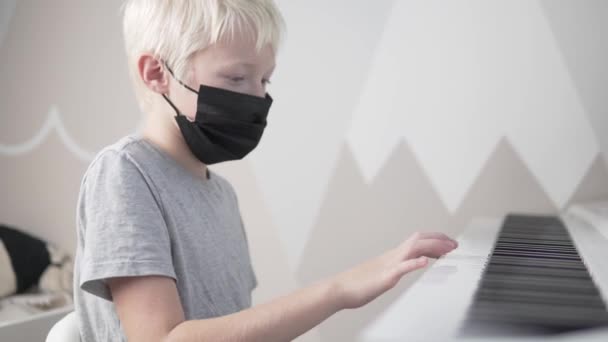 Un chico rubio con una máscara facial aprende a tocar el piano en la escuela durante el covid-19 — Vídeo de stock