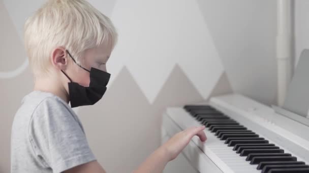 Um menino loiro usando uma máscara médica em seu rosto toca piano na escola durante — Vídeo de Stock