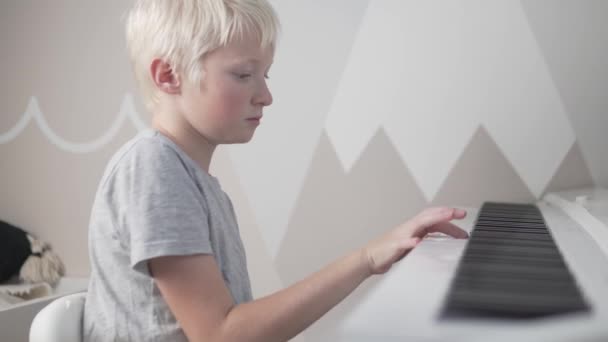 Um menino loiro bonito pratica tocar piano em casa durante a pandemia do covid-19 — Vídeo de Stock