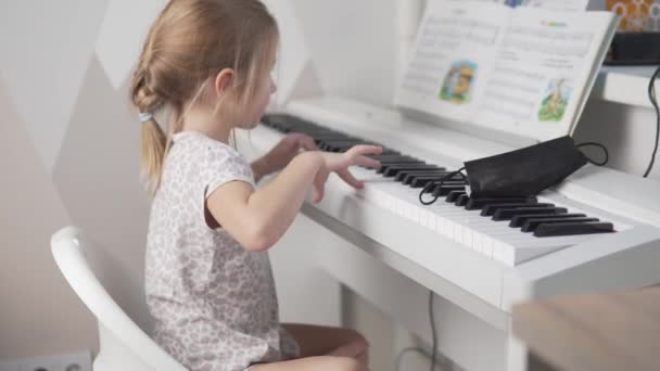 A vista de trás, uma menina brilhante olha para as notas das crianças e toca piano eletrônico — Vídeo de Stock