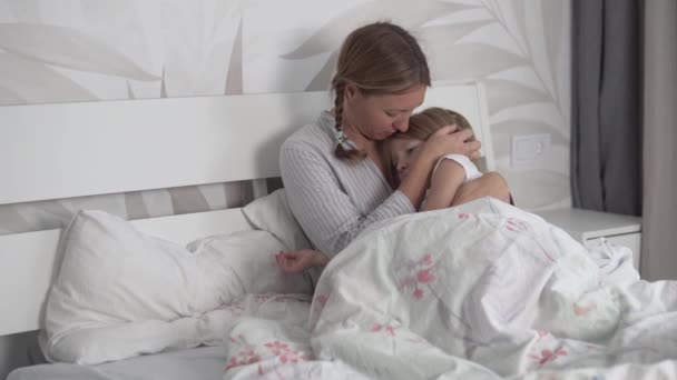 Mama przyciska pięcioletnią córkę do klatki piersiowej i delikatnie głaszcze jej głowę rano w łóżku — Wideo stockowe