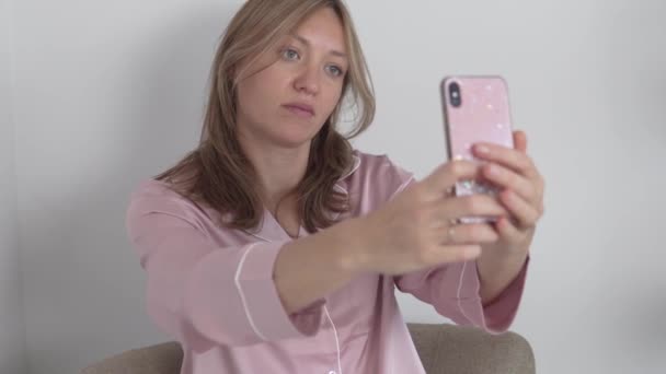Een mooie brunette vrouw in een roze zijden shirt neemt een selfie op haar telefoon thuis — Stockvideo