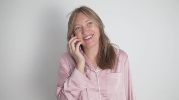 Красива щаслива жінка в шовковій рожевій сорочці говорить на мобільному телефоні вдома — стокове відео