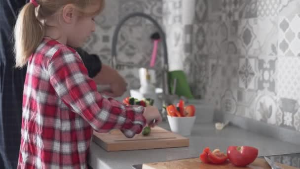 Sevimli küçük bir kız, mutfakta annesiyle birlikte tahta kesme tahtasının üzerinde salatalıkları pipetlere ayırıyor. — Stok video