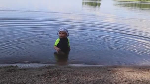 Egy kisfiú kalapban ül a tóban nyáron, és vizet fröcsköl. — Stock videók