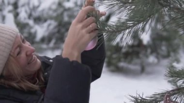 A happy woman hangs a pink ball on a Christmas tree in the forest in winter in Russia