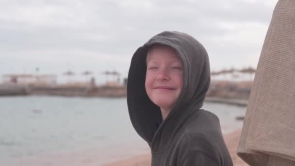 Przystojny blond chłopiec zdejmuje kaptur z głowy na tle morza i uśmiecha się do kamery — Wideo stockowe
