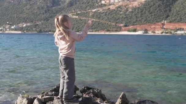 Mała dziewczynka stoi na skałach nad morzem i macha ręką — Wideo stockowe