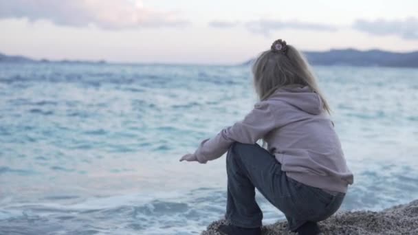 Un bambino si siede su una spiaggia di ciottoli vicino al mare — Video Stock