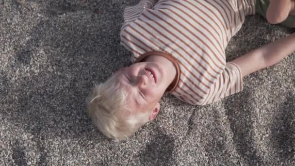 En blond pojke ligger på stranden och skyller på solen — Stockvideo