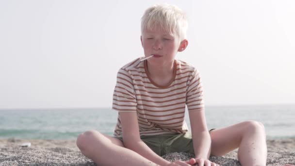 Egy szőke fiú nyalókát szopogat a tengerparton. — Stock videók