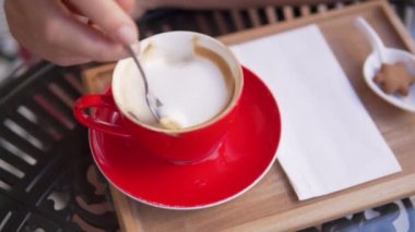Yakın plan, ahşap bir gözenekte bir bardak kapuçino. Kadınların eli kaşık köpüklü latte köpüğünü temizler.