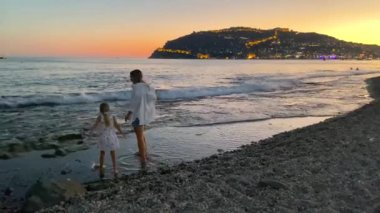 Anne ve küçük kızı yazın gün batımında deniz kenarında yürürler. Türkiye, Alanya