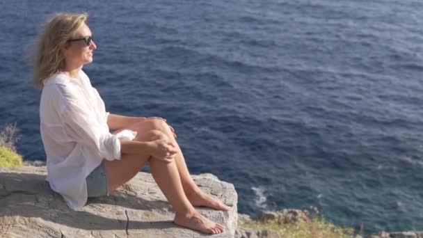美しい女性は海のそばの岩の上に座って景色を楽しむ — ストック動画