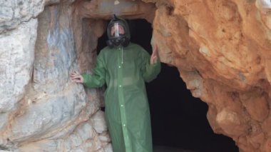 Gaz maskeli ve koruyucu pelerinli bir kadın karanlık bir mağaradan aydınlığa çıkıyor. Kıyamet