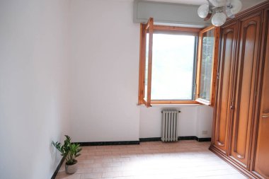 Beyaz duvarlı oda, açık pencere, ahşap gardırop, avize ve duvarın karşısında bir bitki. İç mimari